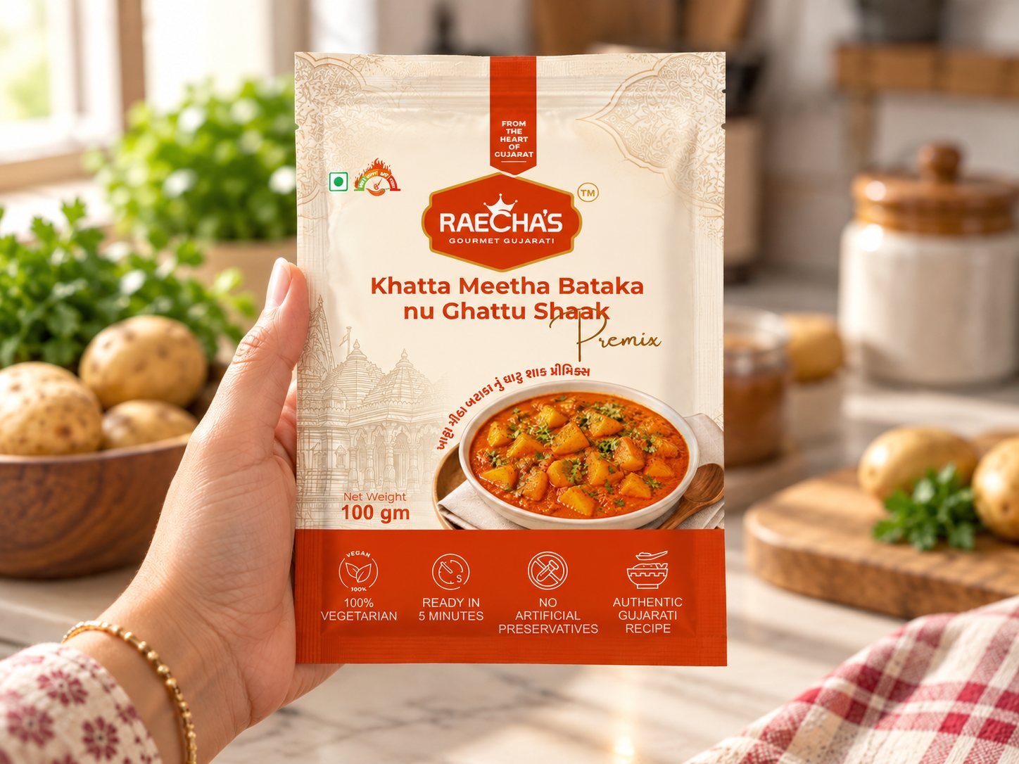 A hand holding a Raecha's Khatta Meetha Bataka nu Ghattu Shaak Premix pouch in a kitchen, with potatoes on the counter.